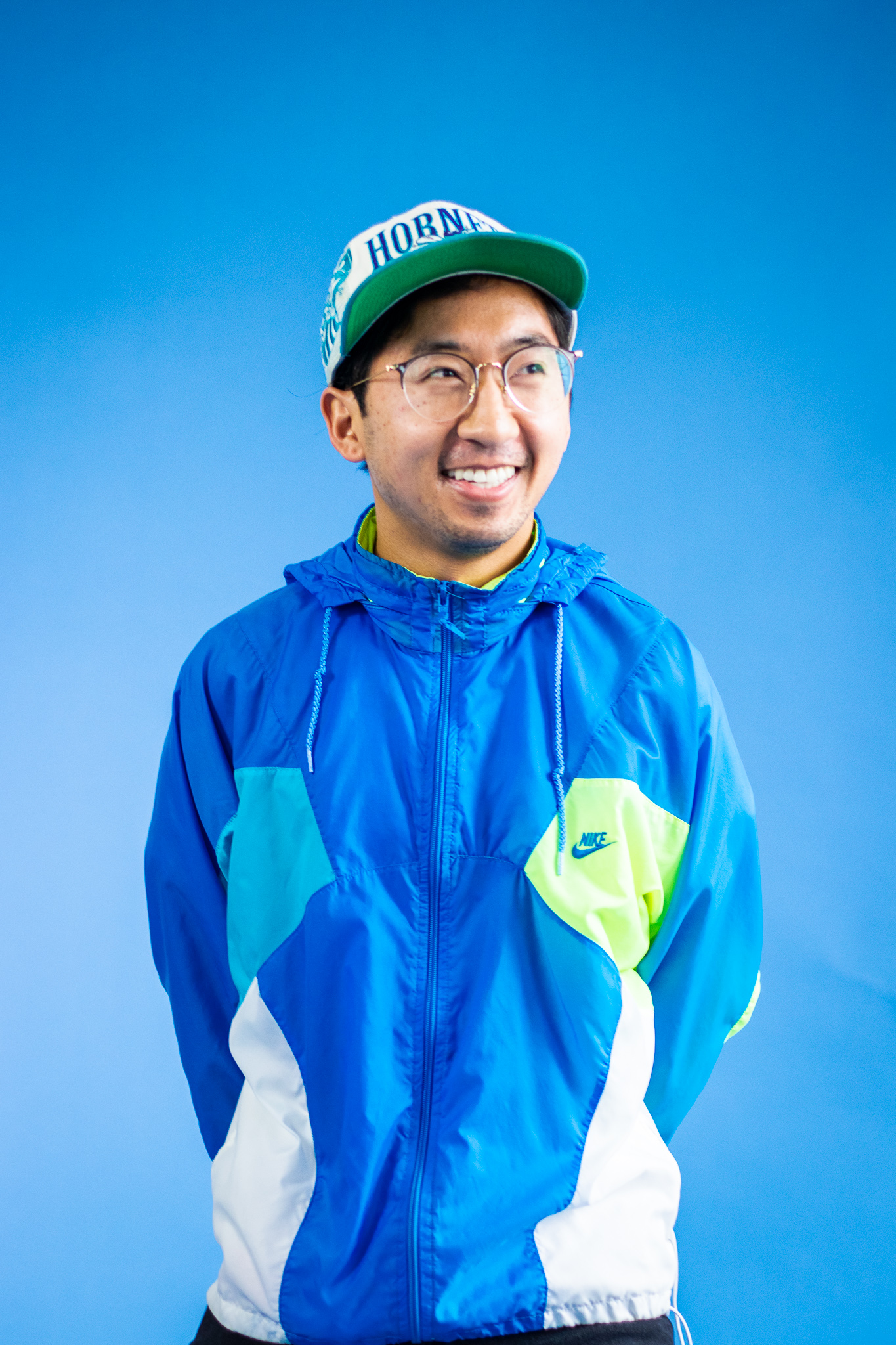 Francis Orallo, product designer, wearing a blue Nike windbreaker and green cap, smiling warmly against a vibrant blue background